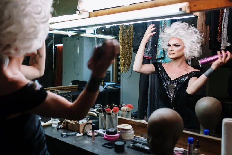 Performer spraying a platinum wig in front of a bulb-lit dressing-room mirror