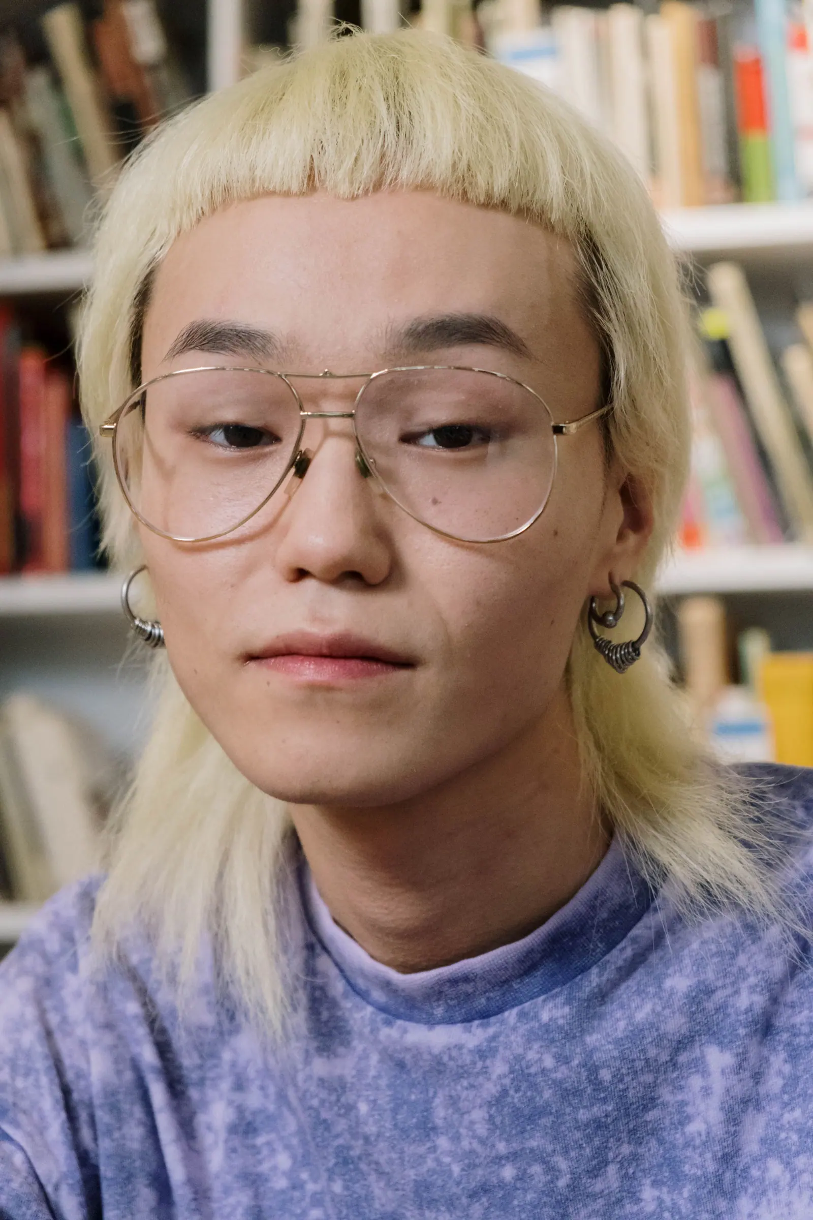 Person with a platinum blonde mullet and round glasses in front of a bookshelf