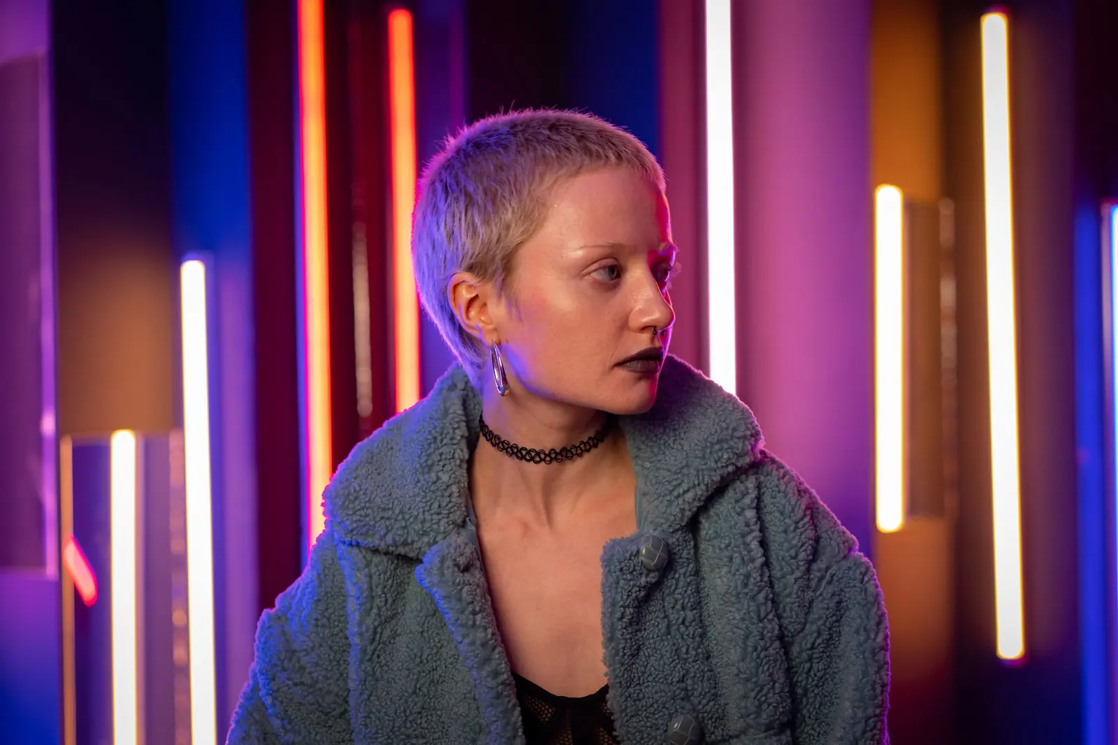 Person with a platinum buzz cut in a fleece jacket, lit by violet and amber neon tubes