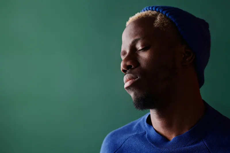 Serene portrait of a man with closed eyes and a blue hat, against a green background, expressing calm and relaxation.