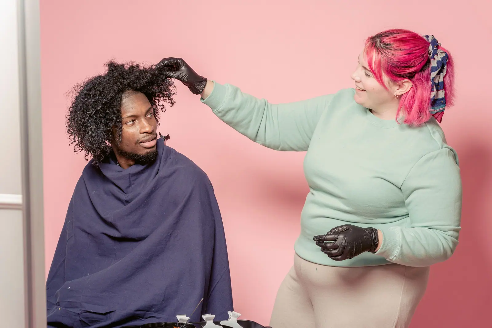 Pink-haired stylist in gloves shaping a client's big curls against a bubblegum-pink wall