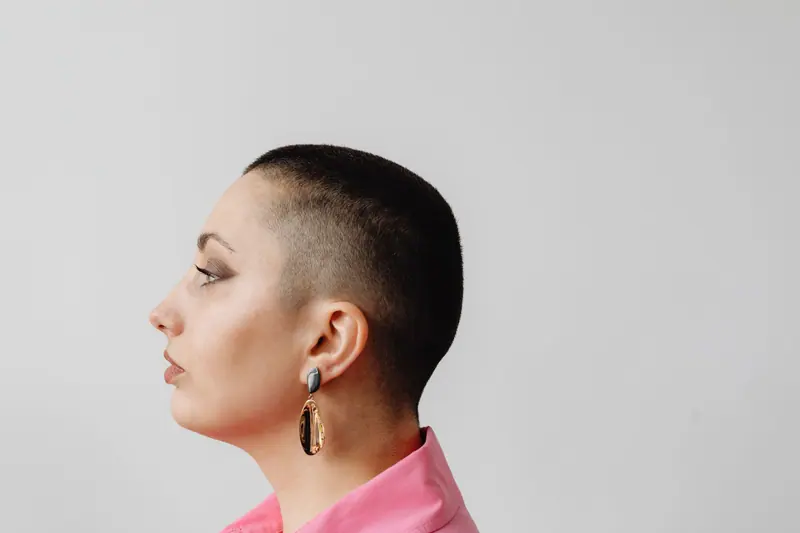 Profile view of a woman with a buzzcut wearing an earring, featuring a neutral background.