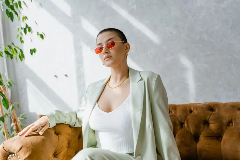 Confident woman in red sunglasses and chic suit sitting on a vintage couch in a sunlit room.
