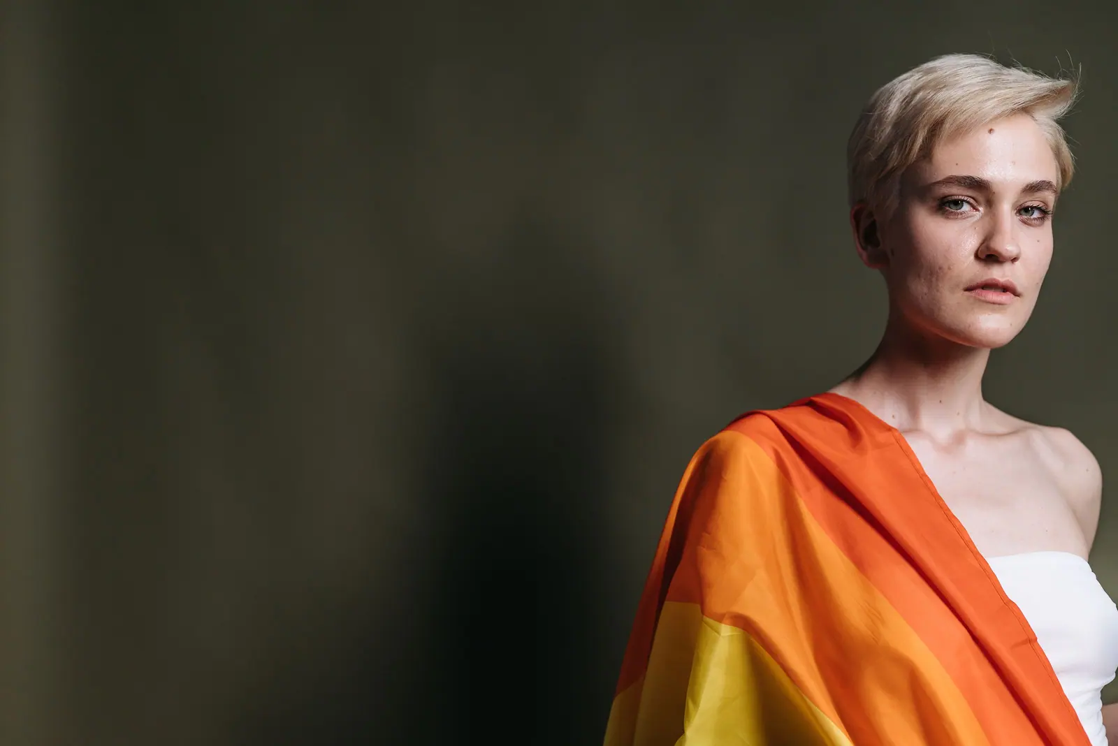 Woman with a cropped platinum cut wearing a rainbow pride flag over her shoulder against a dark wall