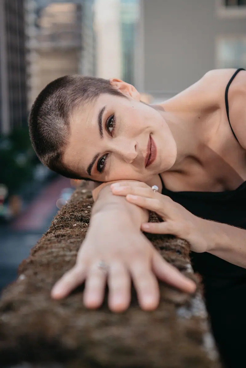 Close-up portrait of a serene woman with a buzz cut, arm extended, smiling gently.