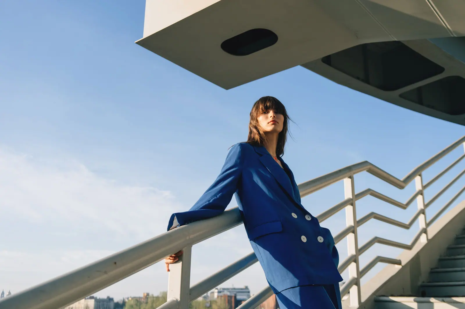 Person in a cobalt double-breasted suit leaning on a white bridge railing against a bright blue sky