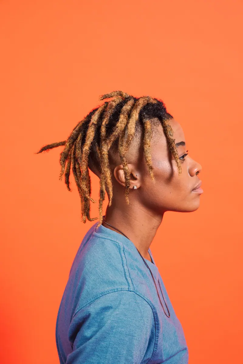 Stylish profile portrait of an individual with dreadlocks against a bright orange background.