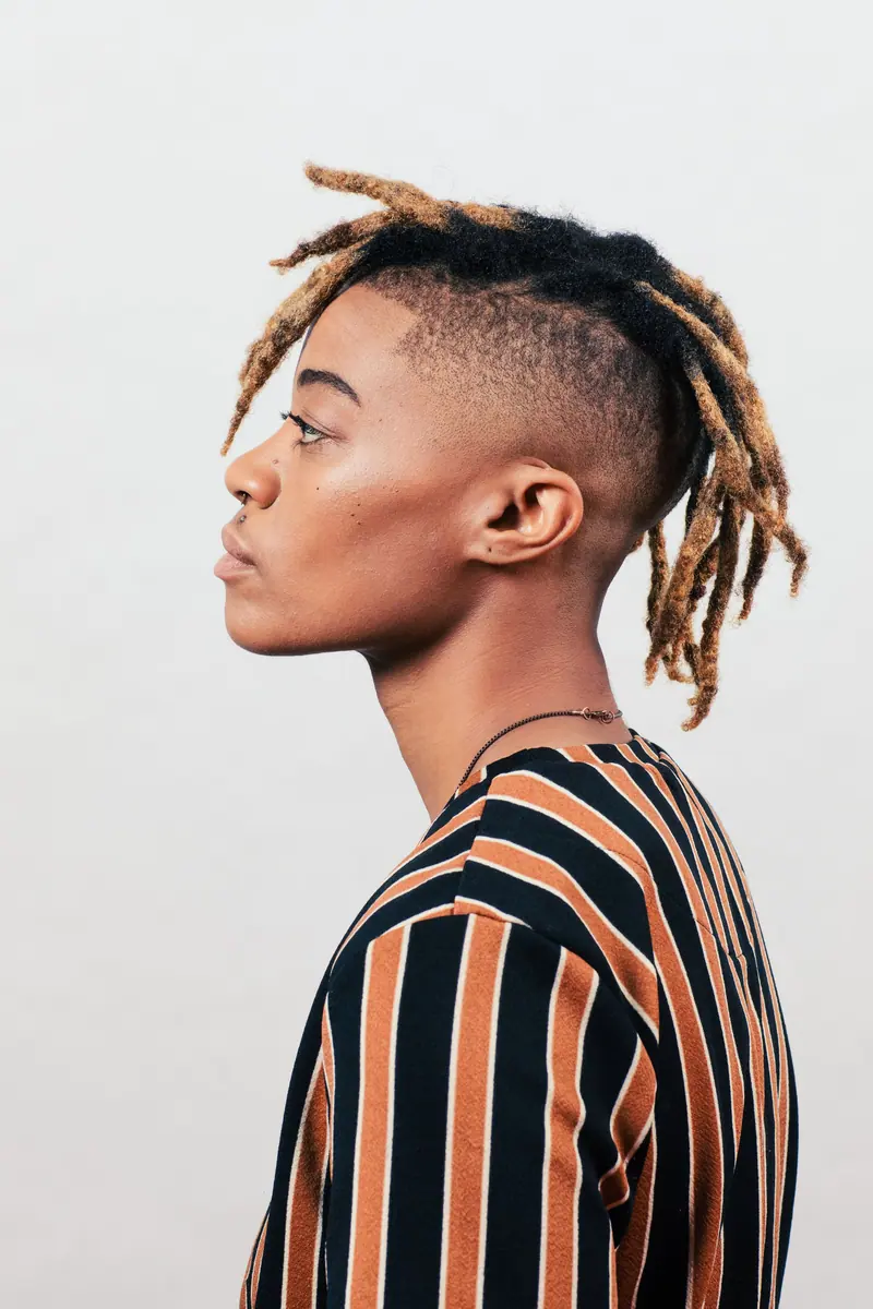 Profile portrait of a young person with distinctive loc hairstyle wearing a striped shirt against a white background.