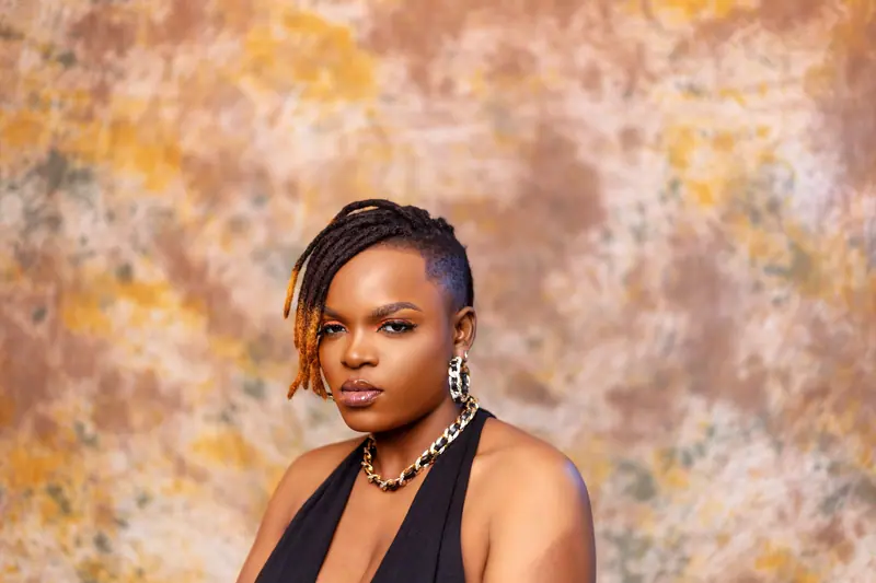 A striking portrait of a confident woman with dreadlocks and bold jewelry against a textured backdrop.