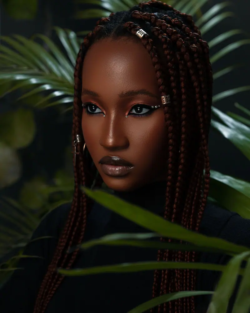 Striking portrait of a woman with box braids, surrounded by lush foliage.