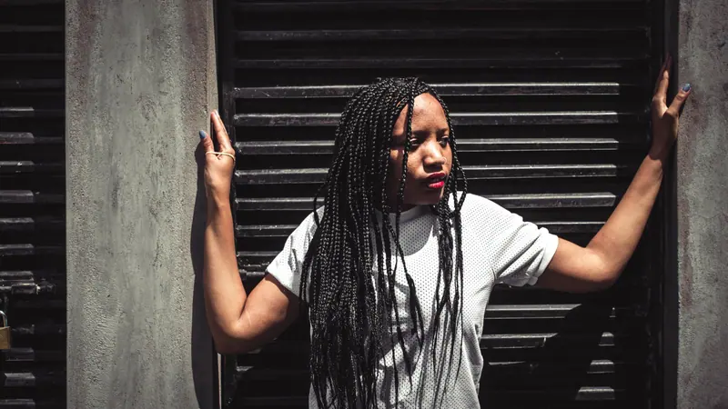 Stylish woman with braided hair posing outdoors against a black vent wall, exuding confidence.