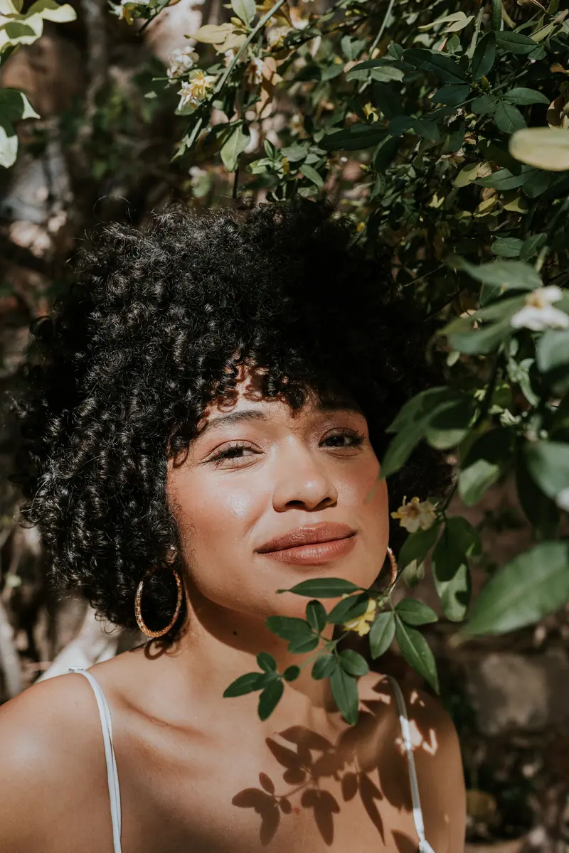A joyful woman with curly hair posing amidst lush greenery on a sunny day outdoors.