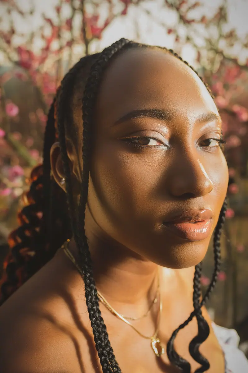 Portrait of a woman with braided hair, bathed in sunlight, among blossoming trees.