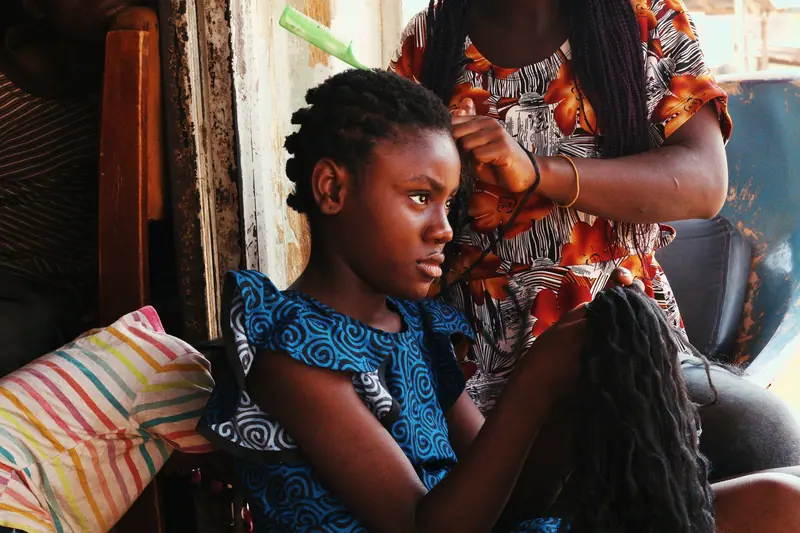 A girl gets her hair braided at home, showcasing cultural hair styling.