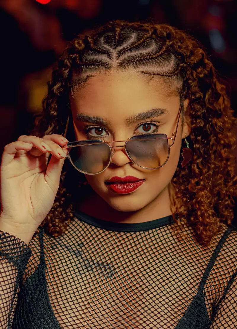 Fashion portrait of a woman wearing cornrow braids, sunglasses, and a fishnet top, exuding confidence.