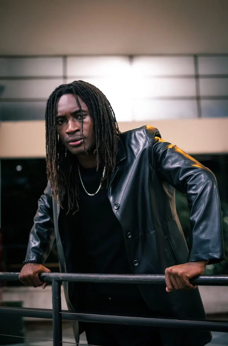 Portrait of a fashionable young man with locs, wearing a black coat, leaning on a railing at night.