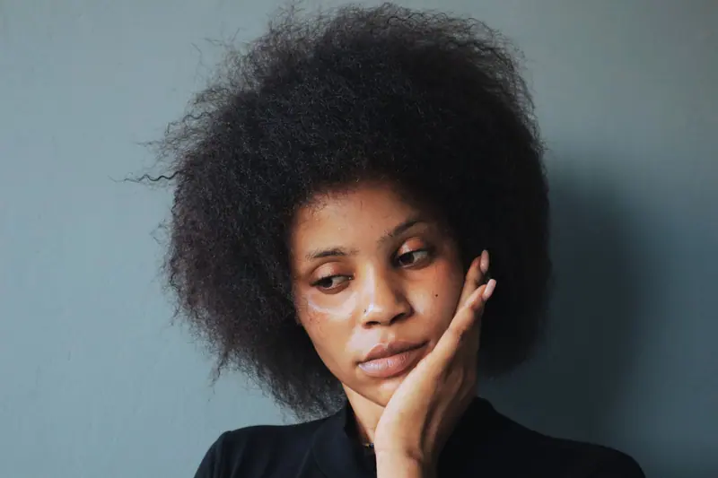 Woman with a full natural afro resting her chin in her hand against a blue-grey wall