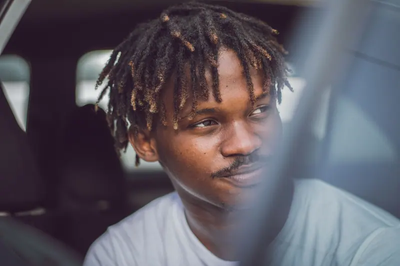 Close-up portrait of a young man with dreadlocks sitting in a car, looking thoughtful.