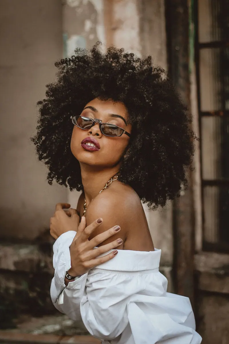 Elegant fashion portrait of a woman with curly afro hair, stylish sunglasses, and white blouse.