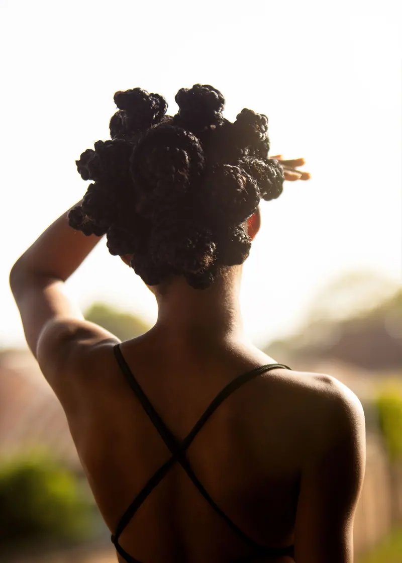 A woman with intricately braided hair stands outdoors, facing the sunlight.