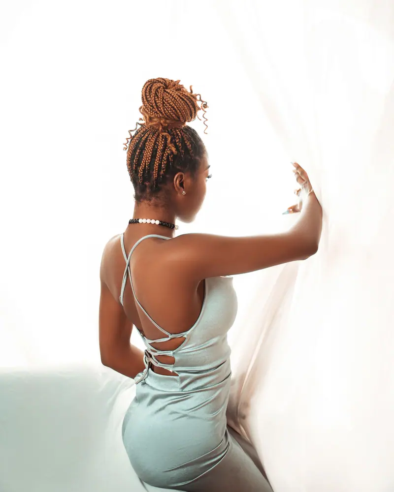 Elegant woman in stylish dress with braided updo looking out a bright window.