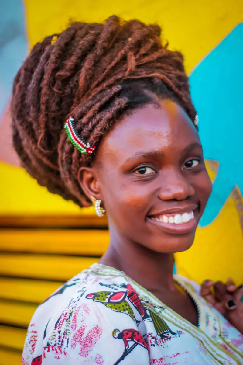A cheerful woman with intricate dreadlocks wearing vibrant clothing against a vivid background.