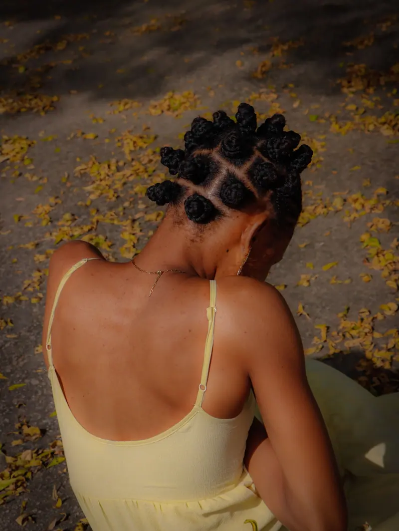 A woman sitting outdoors in a yellow dress, showcasing bantu knots hairstyle during daytime.