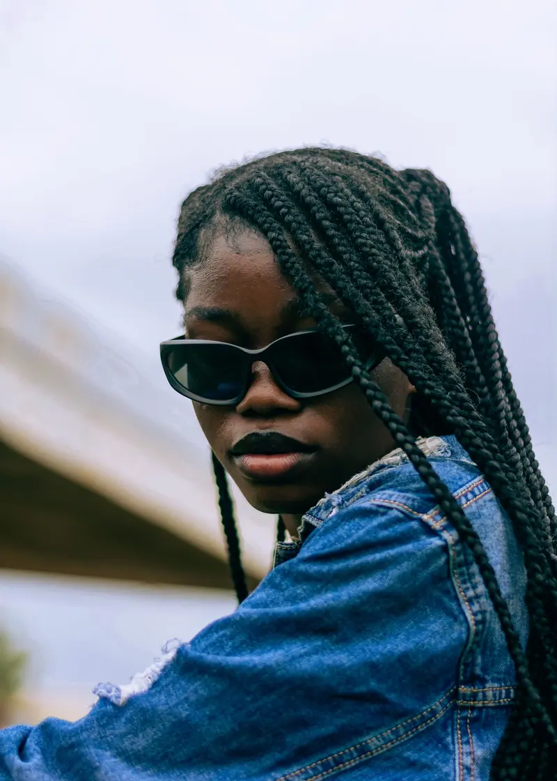 Fashionable young woman with braids and sunglasses wearing a denim jacket outdoors.