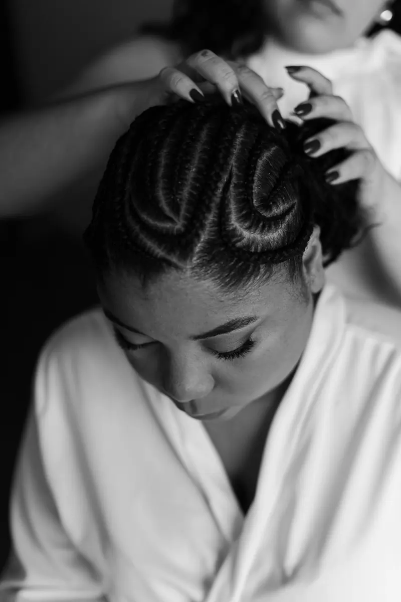 Black and white photo capturing a bride's intricate braided hairstyle being styled, highlighting bridal preparation.