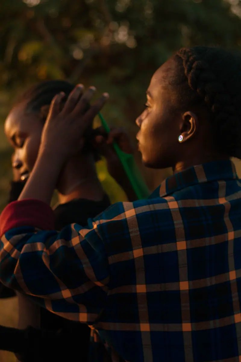 Two women braiding hair in warm sunset light, outdoor setting.