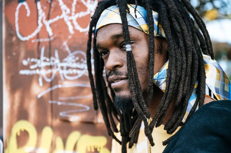 Close-up portrait of a man with dreadlocks and a colorful headscarf, captured outdoors with graffiti background.