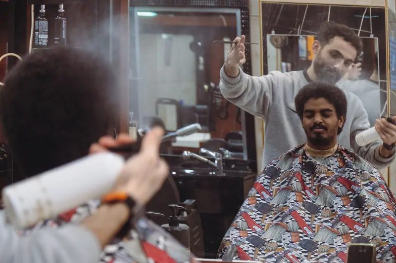 Barber styling hair of customer with spray and tools in a modern barbershop setting.