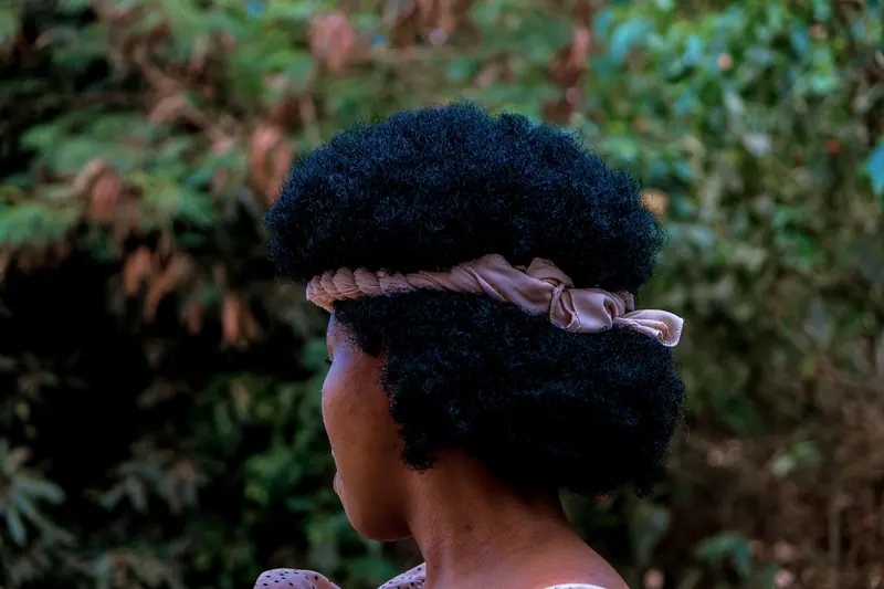 A woman with a beautiful natural afro and headband standing outdoors among greenery.