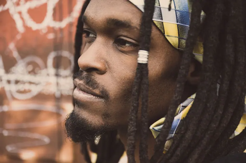A thoughtful man with dreadlocks and a headband stands outdoors, exuding style and individuality.