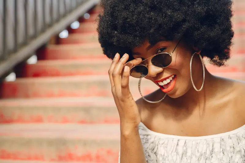 Charming woman with afro hairstyle and sunglasses smiling on colorful stairs.