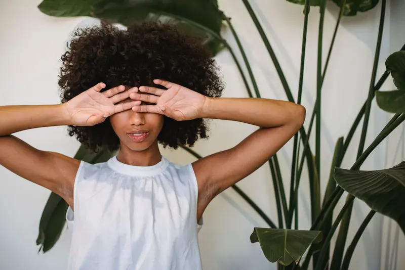 A young woman in a white blouse with curly hair covering her eyes, set against a leafy background.