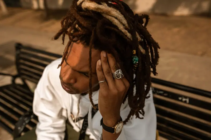A man with dreadlocks looking contemplative while sitting on a bench outdoors.