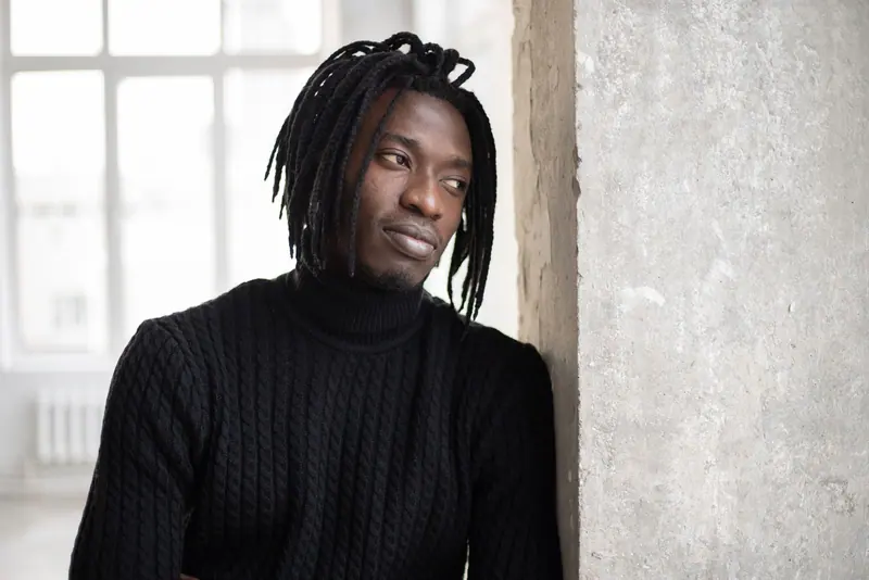 African American man leaning against a concrete wall indoors, looking away pensively.