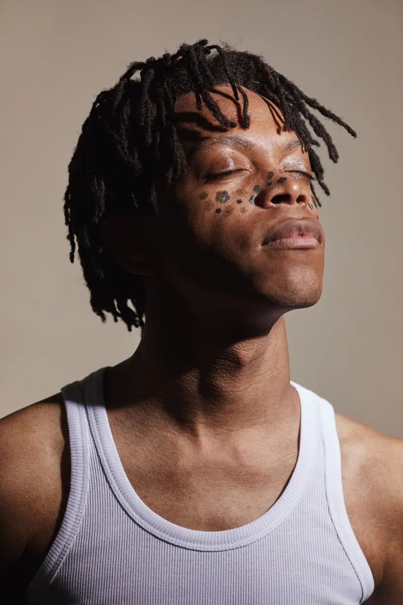 A young man with closed eyes and dreadlocks in a studio setting, wearing a tank top.