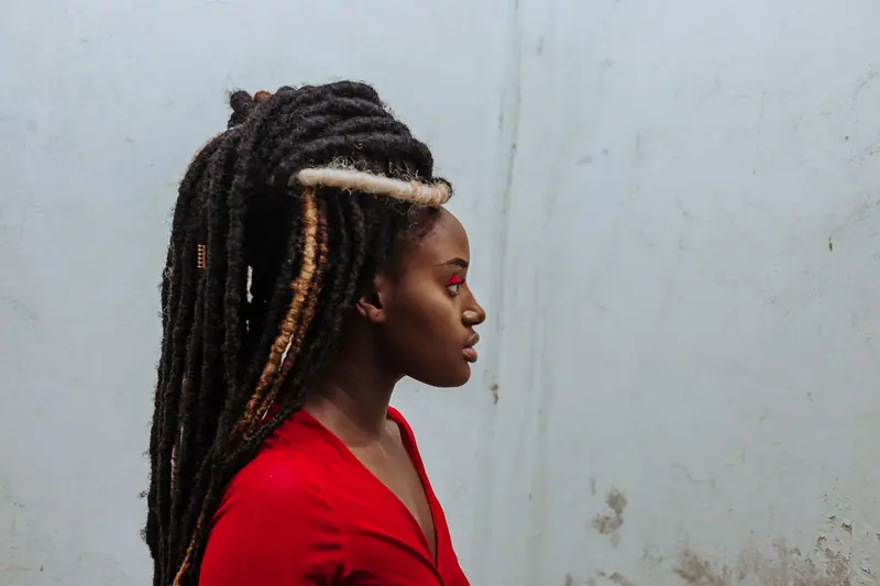 Profile portrait of a woman with dreadlocks in a red outfit against a white wall.
