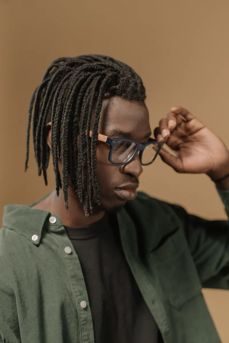 Portrait of a fashionable man with dreadlocks wearing eyeglasses, dressed in a green jacket against a neutral background.