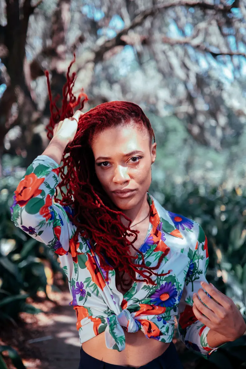 African American woman outdoors touching her red hair, wearing a colorful floral shirt.