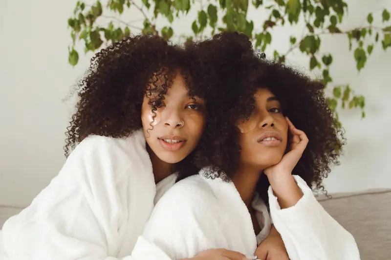 Relaxing portrait of two women with afro hair wearing white bathrobes indoors.