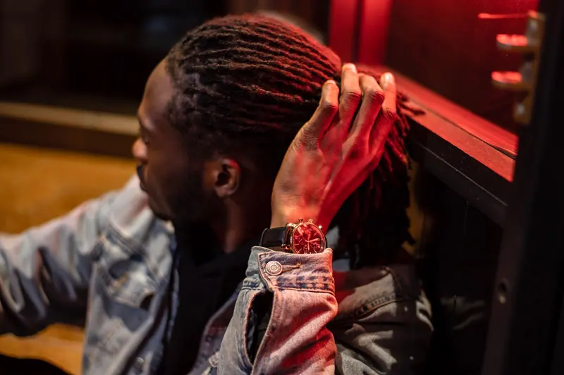 A young man with dreadlocks, in a denim jacket, sits pensively under red neon light.