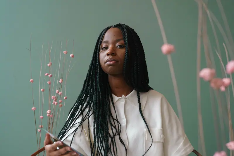 Portrait of a confident young woman with braids, seated indoors with a green backdrop and pink flowers.