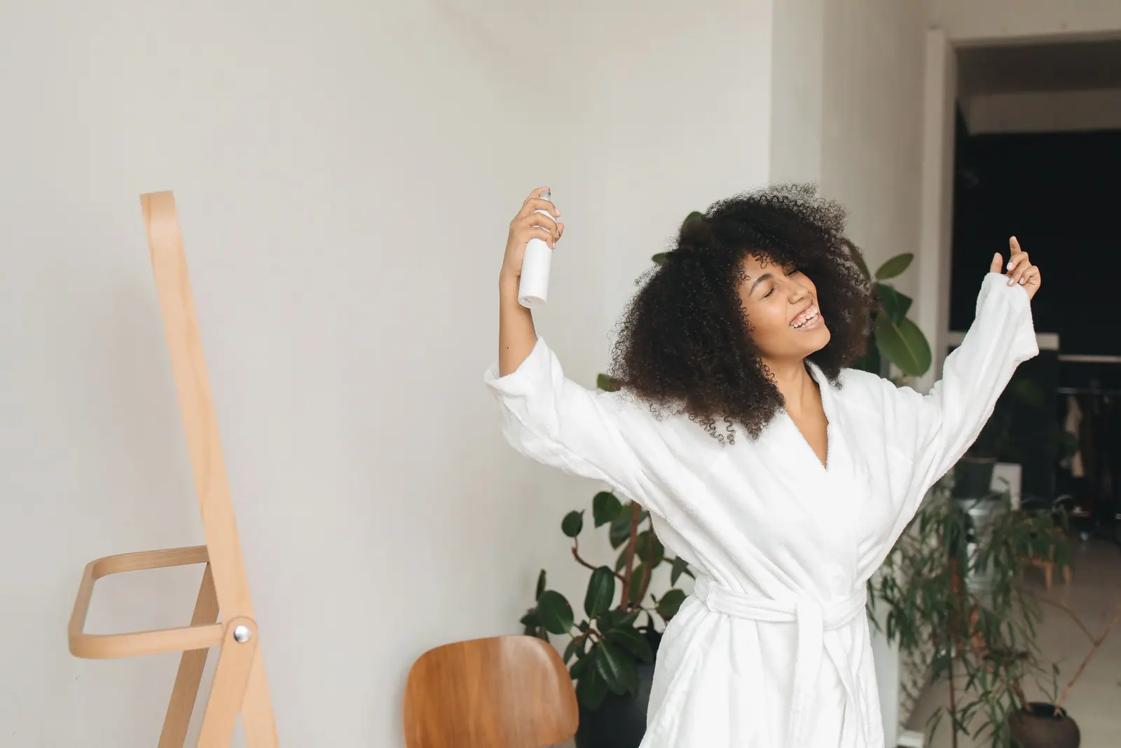 Joyful woman with big natural curls in a white robe spraying leave-in in a sunlit room