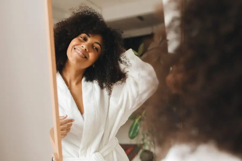 Smiling woman with defined curls checking her hair in a standing mirror