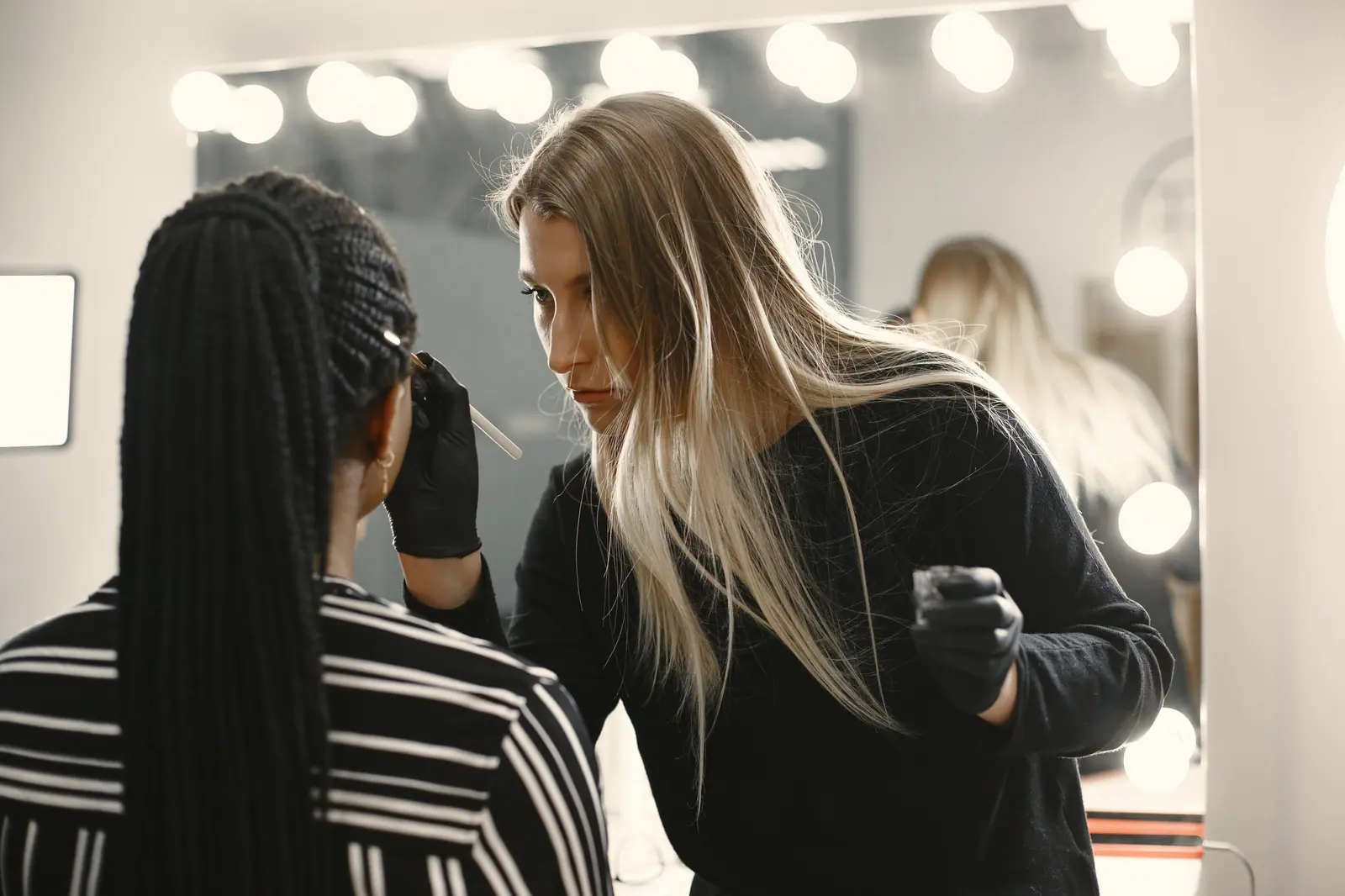 Makeup artist skillfully applies makeup on client in a well-lit studio with mirror lights.