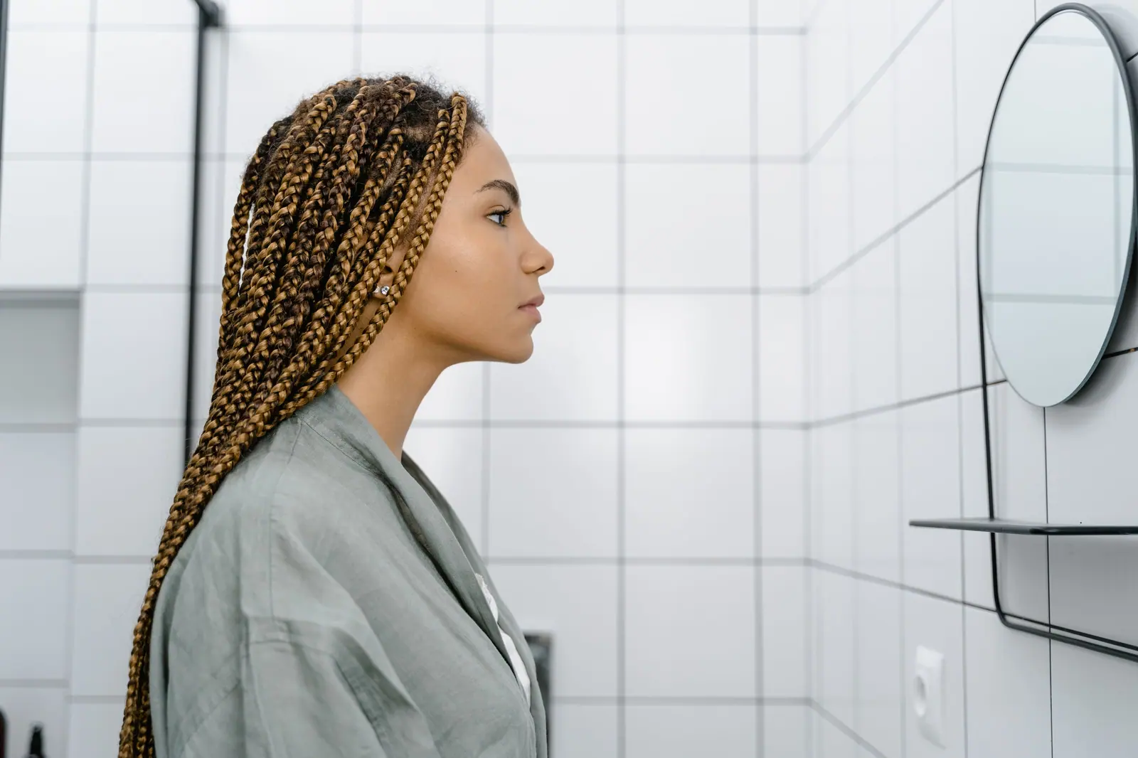 Woman with long honey-brown box braids in profile against a white tiled wall