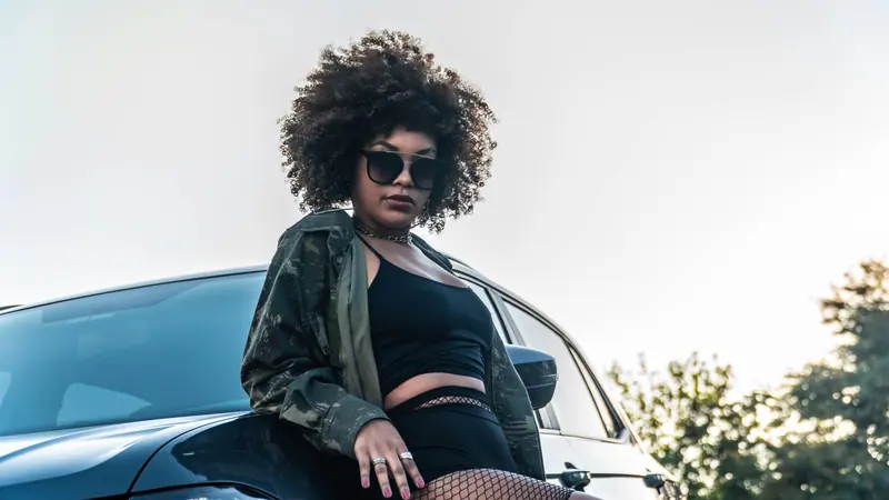 Fashionable young woman with sunglasses and curly hair posing by a sleek car outdoors.